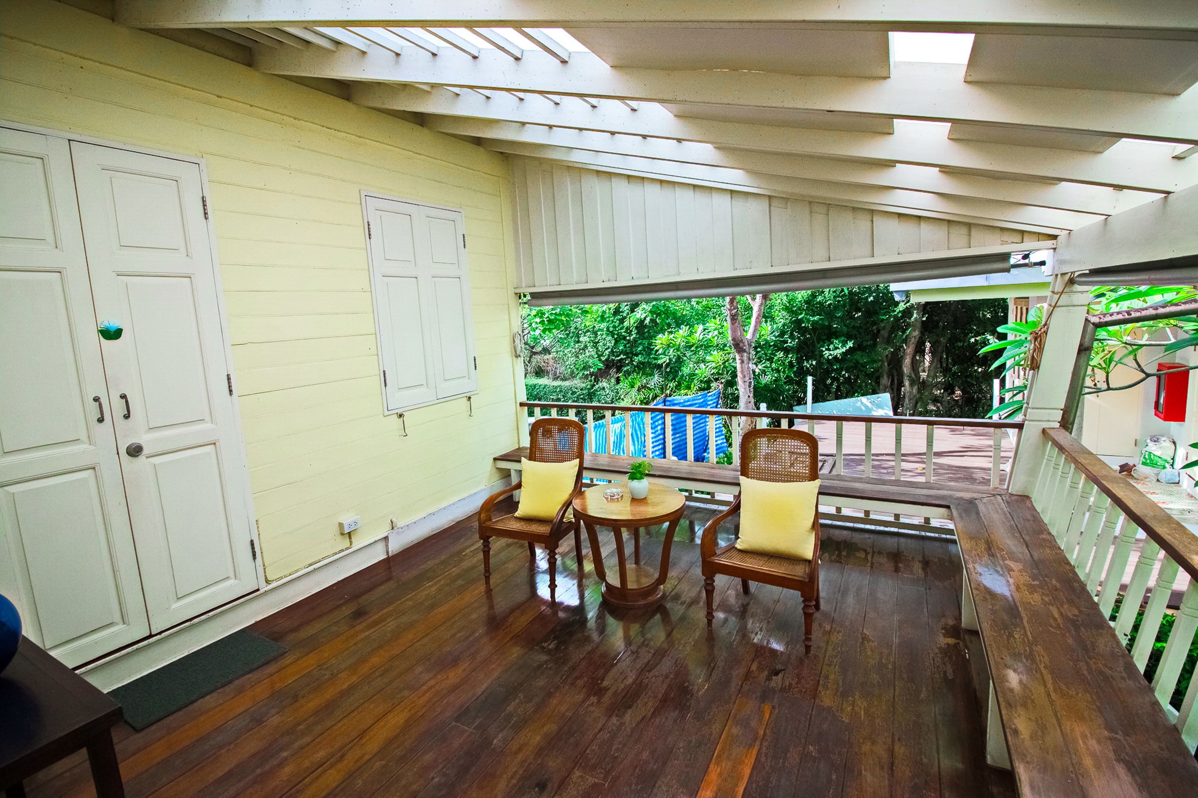 A covered porch with wooden flooring, two wicker chairs, a small round table with a plant, and a bench.