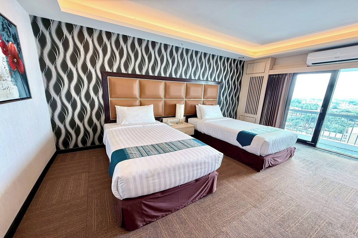 A hotel room with two beds, a patterned wall, a large window, and an air conditioning unit.