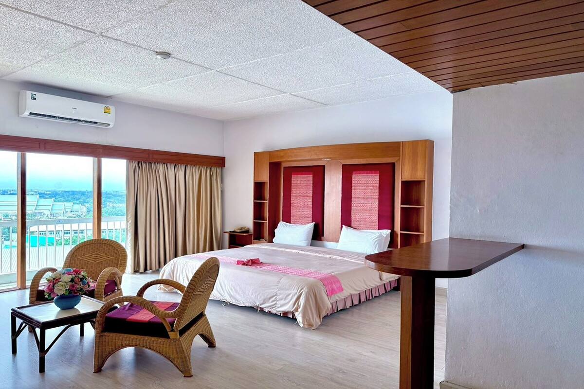 A hotel room with a large bed, a wooden headboard, a dining table, wicker chairs, and a view of the city.