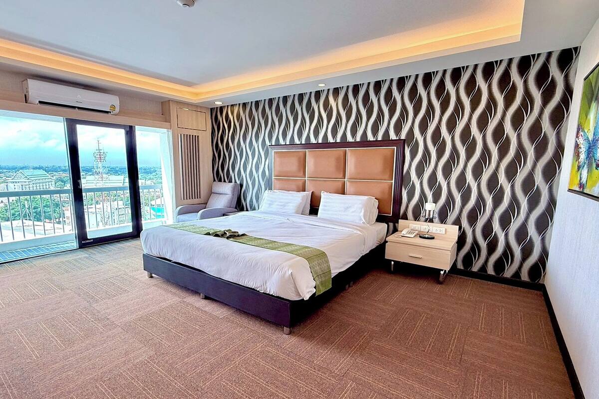 A hotel room with a large bed, a nightstand, a wall-mounted air conditioner, and a view of a cityscape through a large window.