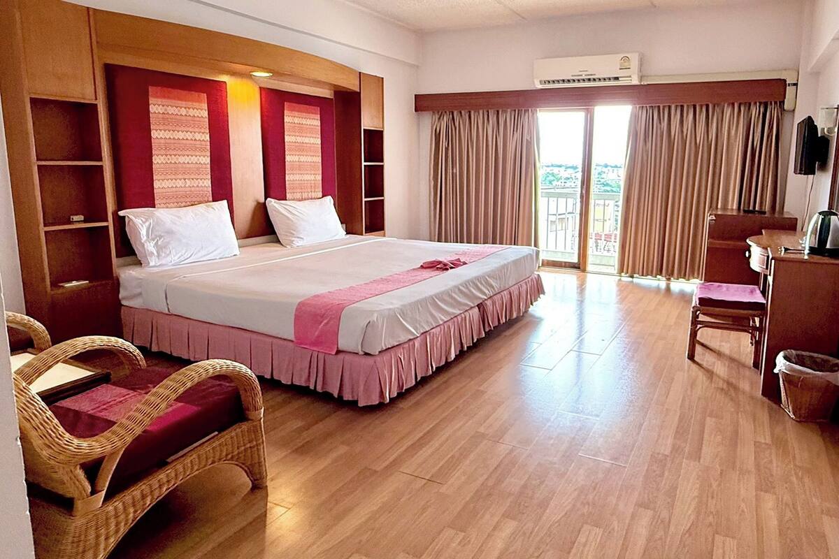 A hotel room with a large bed, a wooden headboard, a desk with a chair, a wicker chair, and a balcony with curtains.