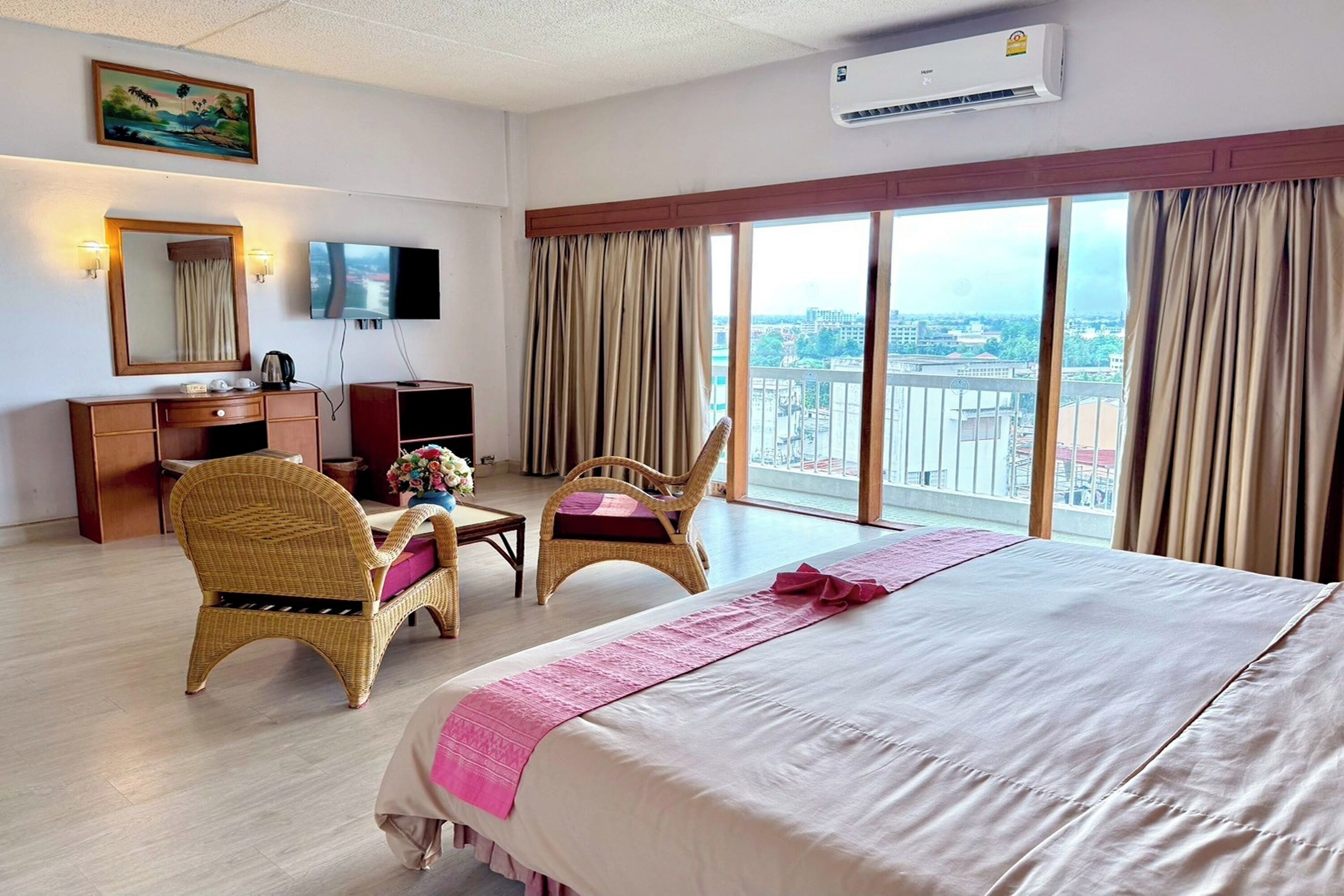A hotel room with a large bed, two wicker chairs, a desk with a television, and a large window with curtains.