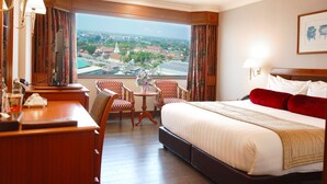 Habitación de hotel con una cama grande, un escritorio, dos sillas, una mesita con un jarrón y vistas a un paisaje urbano a través de amplias ventanas.