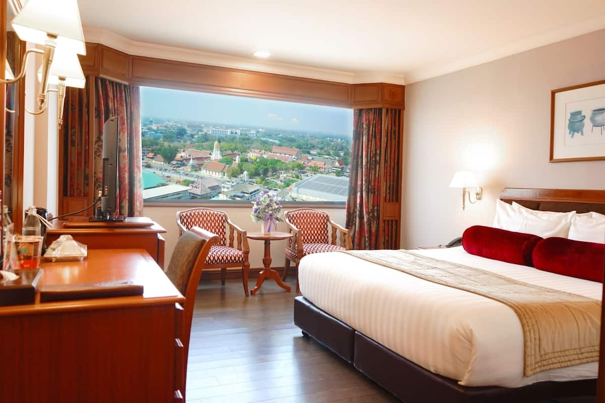 Habitación de hotel con una cama grande, un escritorio, dos sillas, una mesita con un jarrón y vistas a un paisaje urbano a través de amplias ventanas.