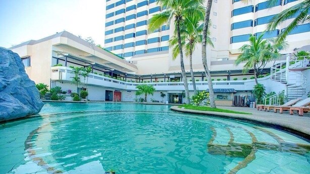 Una piscina con agua cristalina azul, rodeada de palmeras y un edificio moderno con varios pisos y balcones.