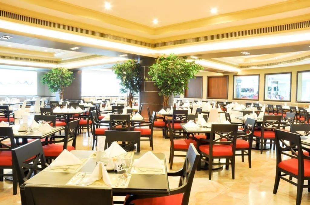 Un comedor amplio con varias mesas y sillas, ventanas grandes y plantas en macetas.
