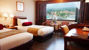 A hotel room with two beds, a desk, a chair, a small table, and a view of a cityscape.