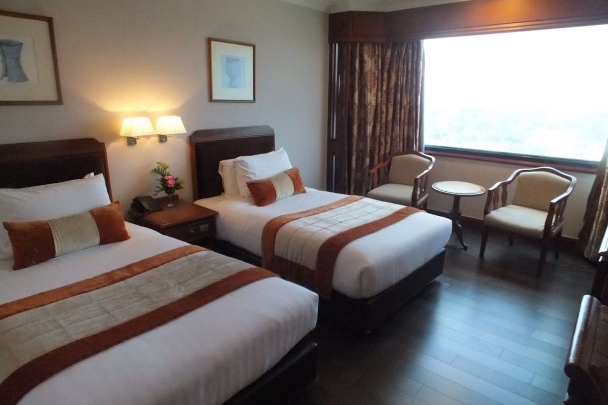 Habitación de hotel con dos camas, una mesita pequeña, dos sillas y un ventanal con cortinas.