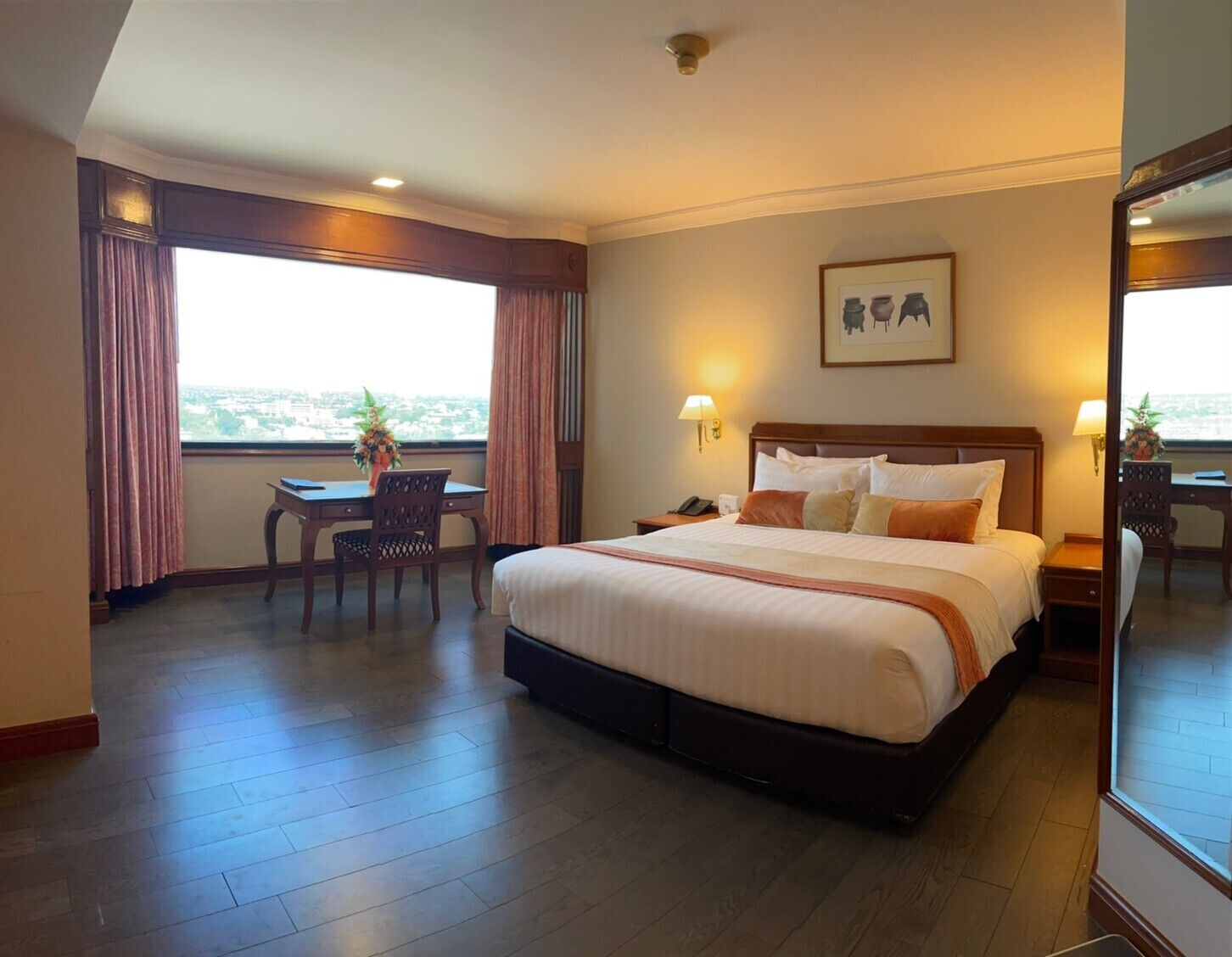 A hotel room with a large bed, a desk with a chair, a window with curtains, and a framed picture on the wall.