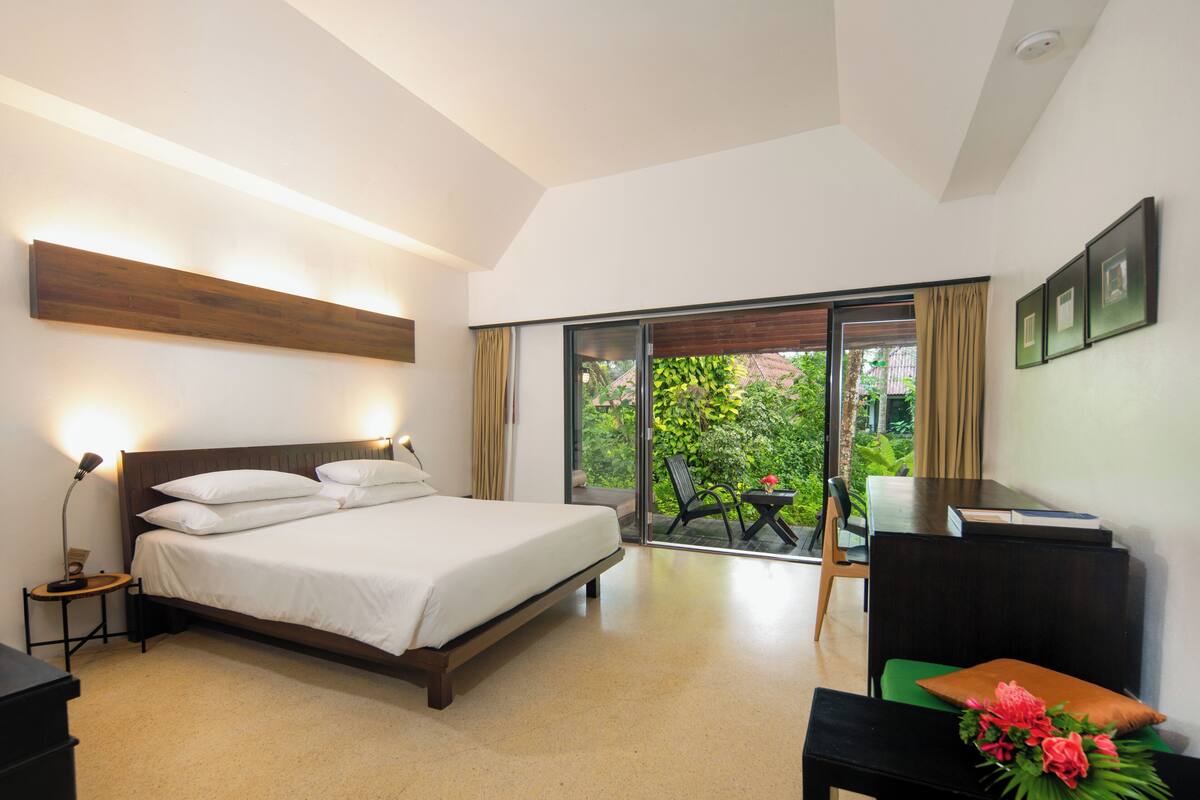 A bedroom with a bed, bedside table, lamp, and a view of greenery through a sliding door.
