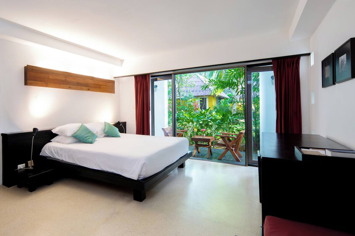 A modern bedroom with a large bed, a desk, and a view of a garden through a sliding door.