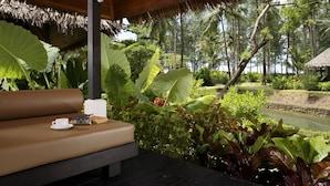 A lounge area with a cushioned bench, a cup, and a book, surrounded by lush tropical plants and a view of a golf course and water body. - Haadson Resort (Takua Pa)