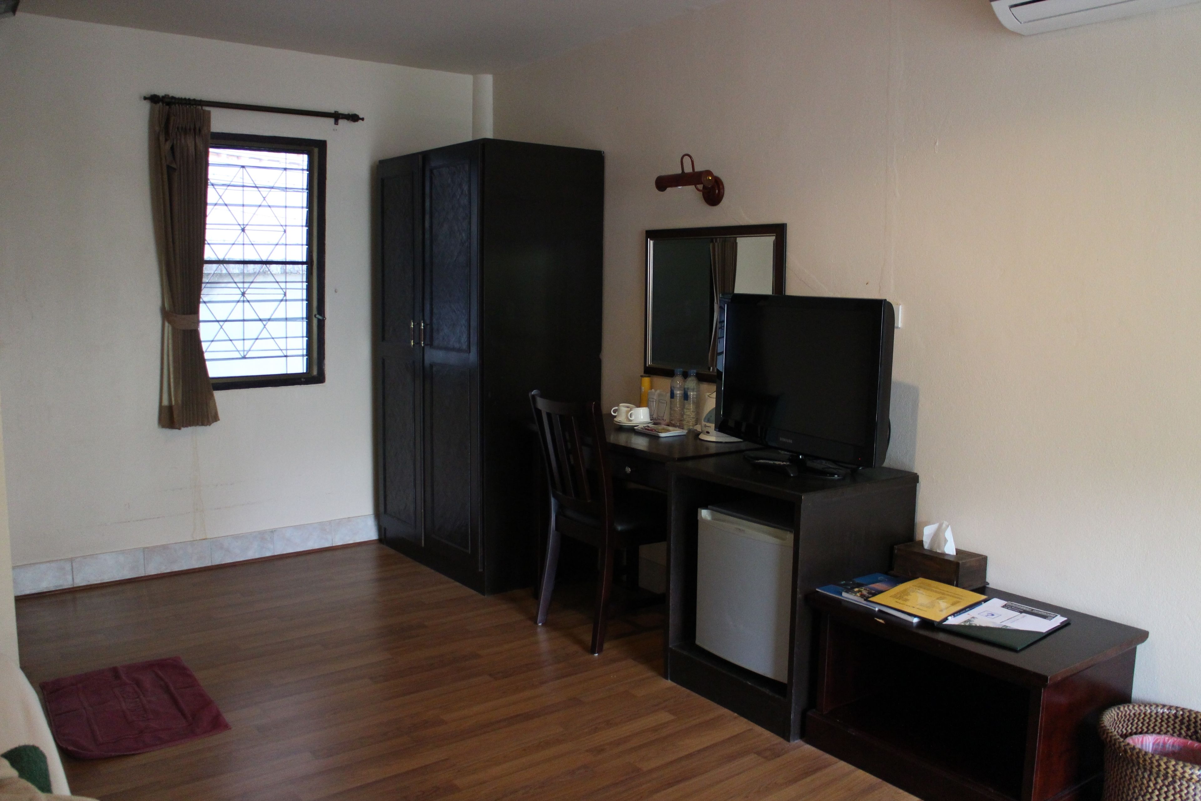 A compact room with a wooden floor, a desk with a computer and a television, a small refrigerator, and a chair.