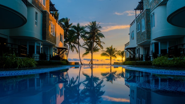 Ein Pool spiegelt Palmen und einen Sonnenuntergang wider, umgeben von Gebäuden mit Balkonen.