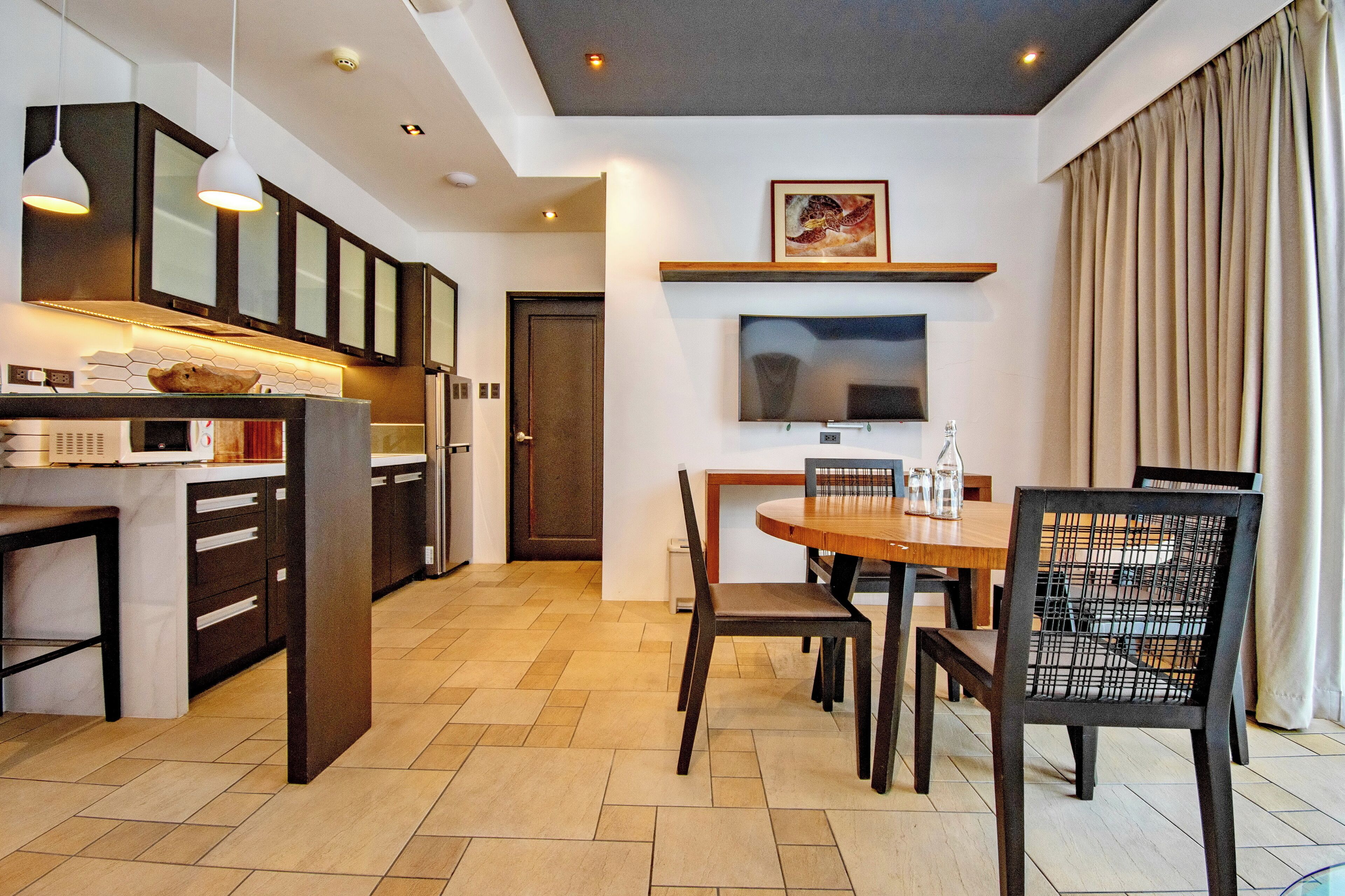 A modern kitchen with dark cabinetry, a dining area with a wooden table and chairs, and a flat-screen TV mounted above the table.