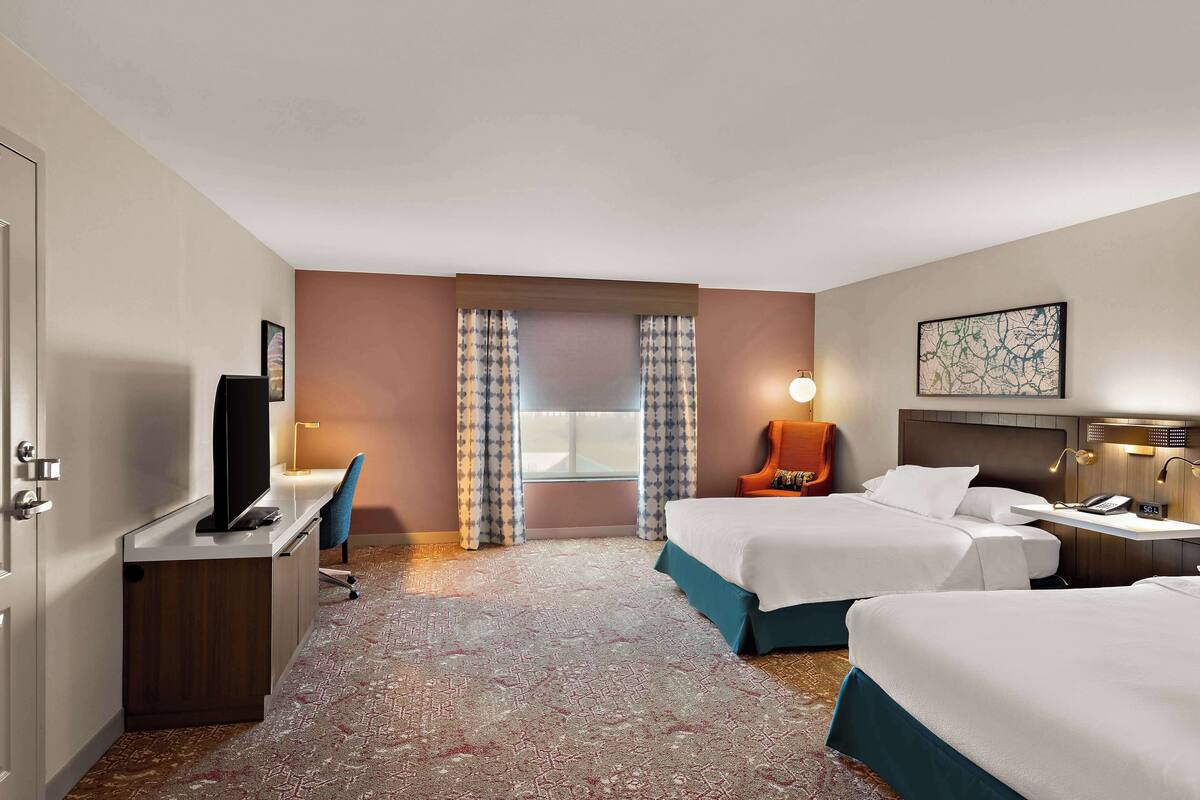 Une chambre d’hôtel avec deux lits, un bureau avec une télévision, une chaise, une lampe de bureau et une fenêtre avec des rideaux.