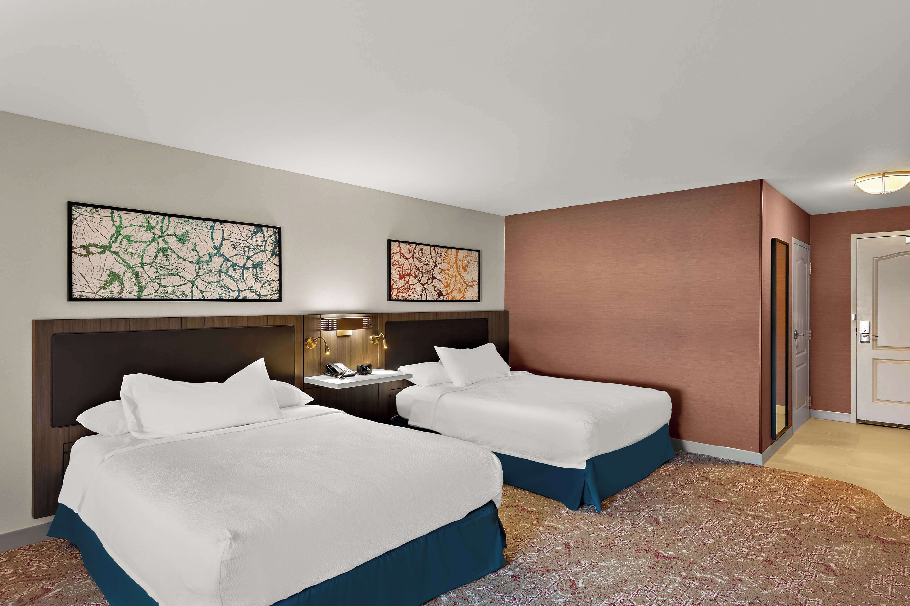 A hotel room with two beds, a nightstand with a lamp, and artwork on the walls.