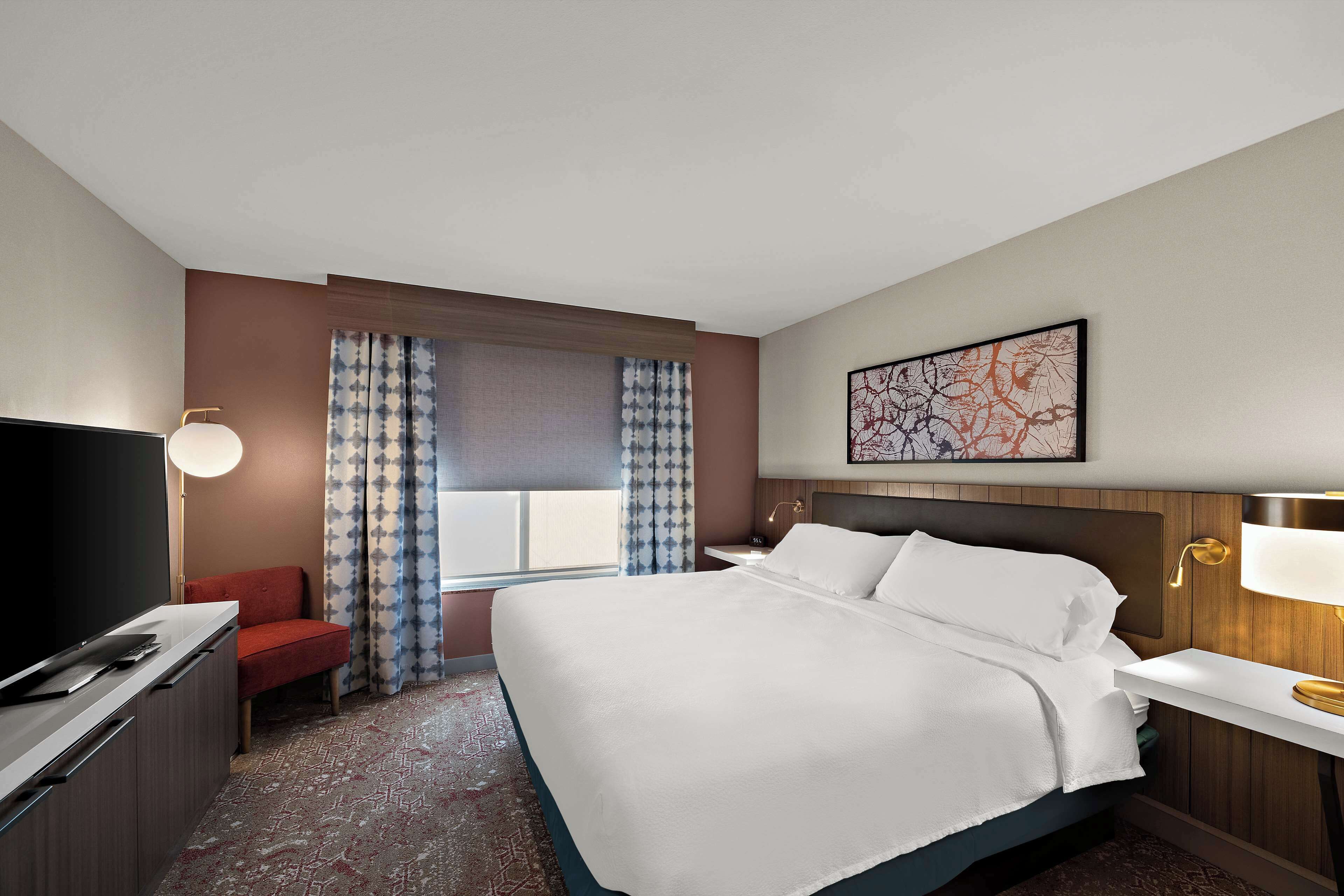 A hotel room with a large bed, a TV, a desk, and a window with curtains.