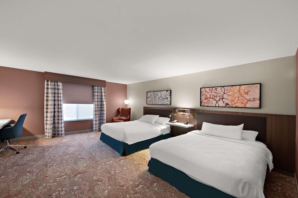 Una habitación de hotel con dos camas, un escritorio, una silla, una mesita de noche con lámpara y obras de arte en las paredes.