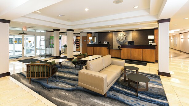 A lobby with a beige sofa, a wooden chair, and a small table. There is a reception desk with a wooden counter and a decorative wall piece. - Fairfield Inn & Suites by Marriott Milwaukee Airport (Oak Creek)