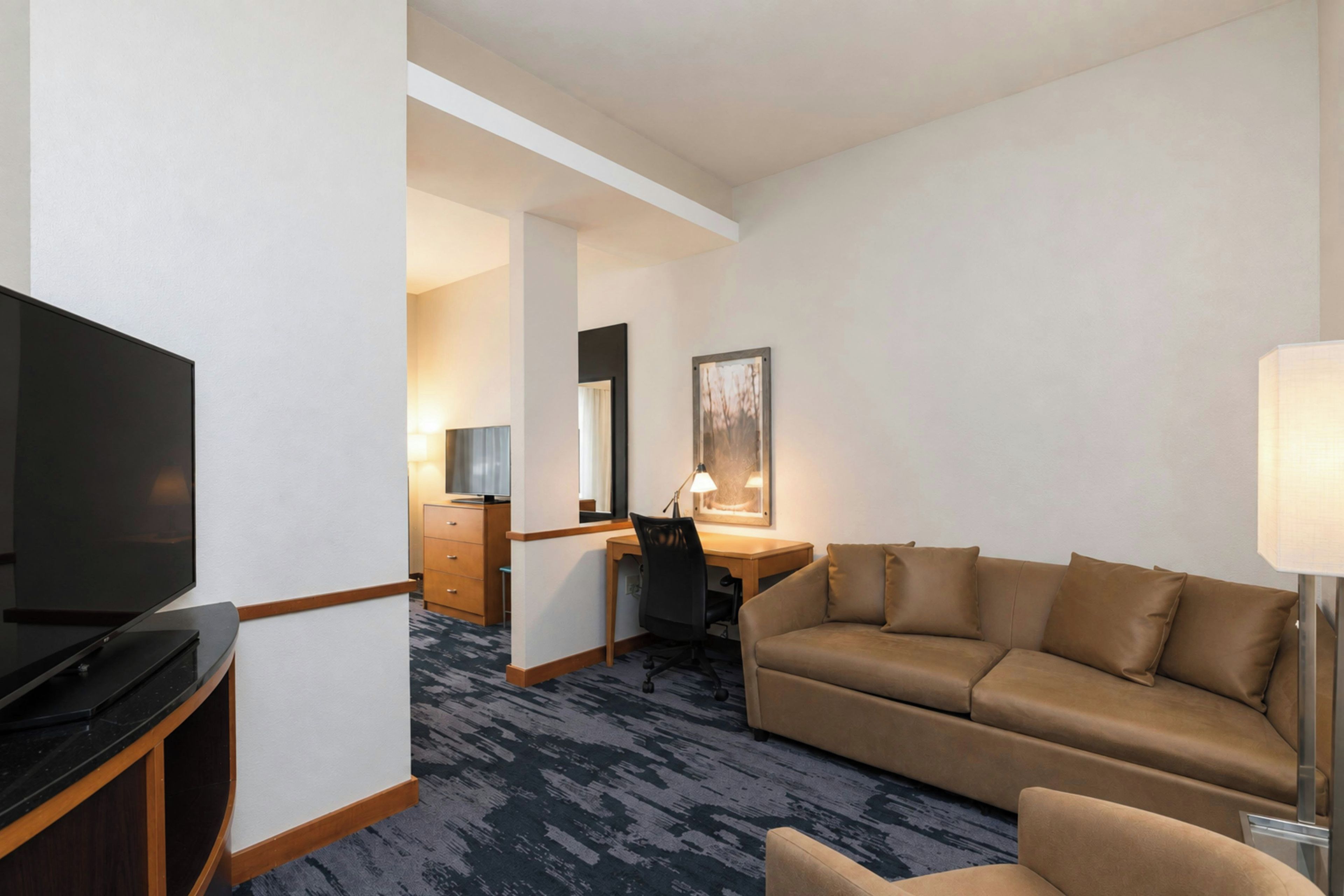 A hotel room with a flat-screen TV, a sofa, a desk with a chair, and a lamp.