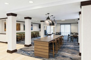 Un moderno vestíbulo de hotel con una zona de bar central, arreglos de asientos y un amplio ventanal que proporciona luz natural.