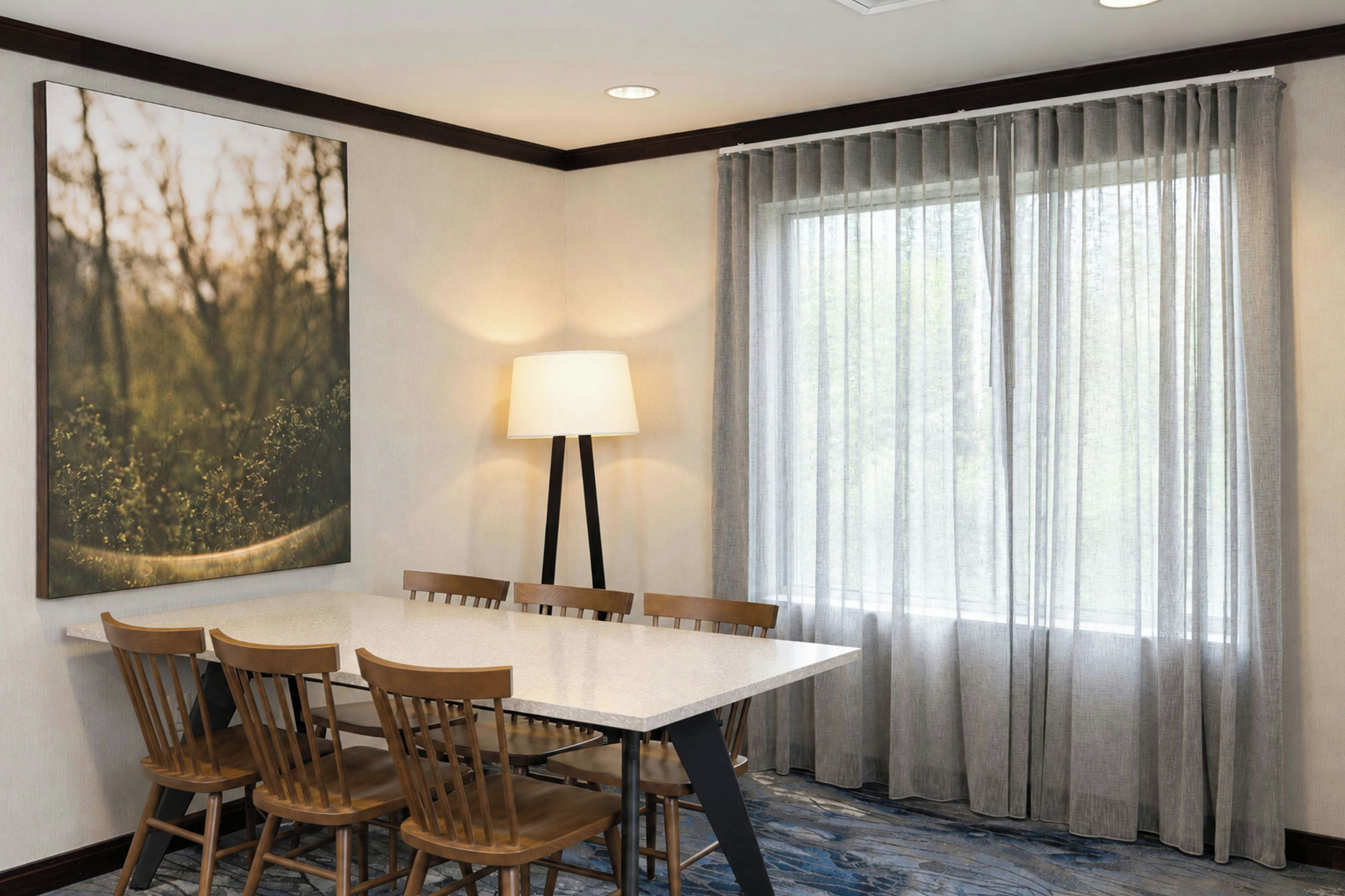 A hotel dining room with a rectangular table, six wooden chairs, a floor lamp, and a large window with sheer curtains.