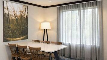 A hotel dining room with a rectangular table, six wooden chairs, a floor lamp, and a large window with sheer curtains.