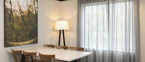 A hotel dining room with a rectangular table, six wooden chairs, a floor lamp, and a large window with sheer curtains.