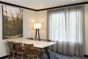 A hotel dining room with a rectangular table, six wooden chairs, a floor lamp, and a large window with sheer curtains.