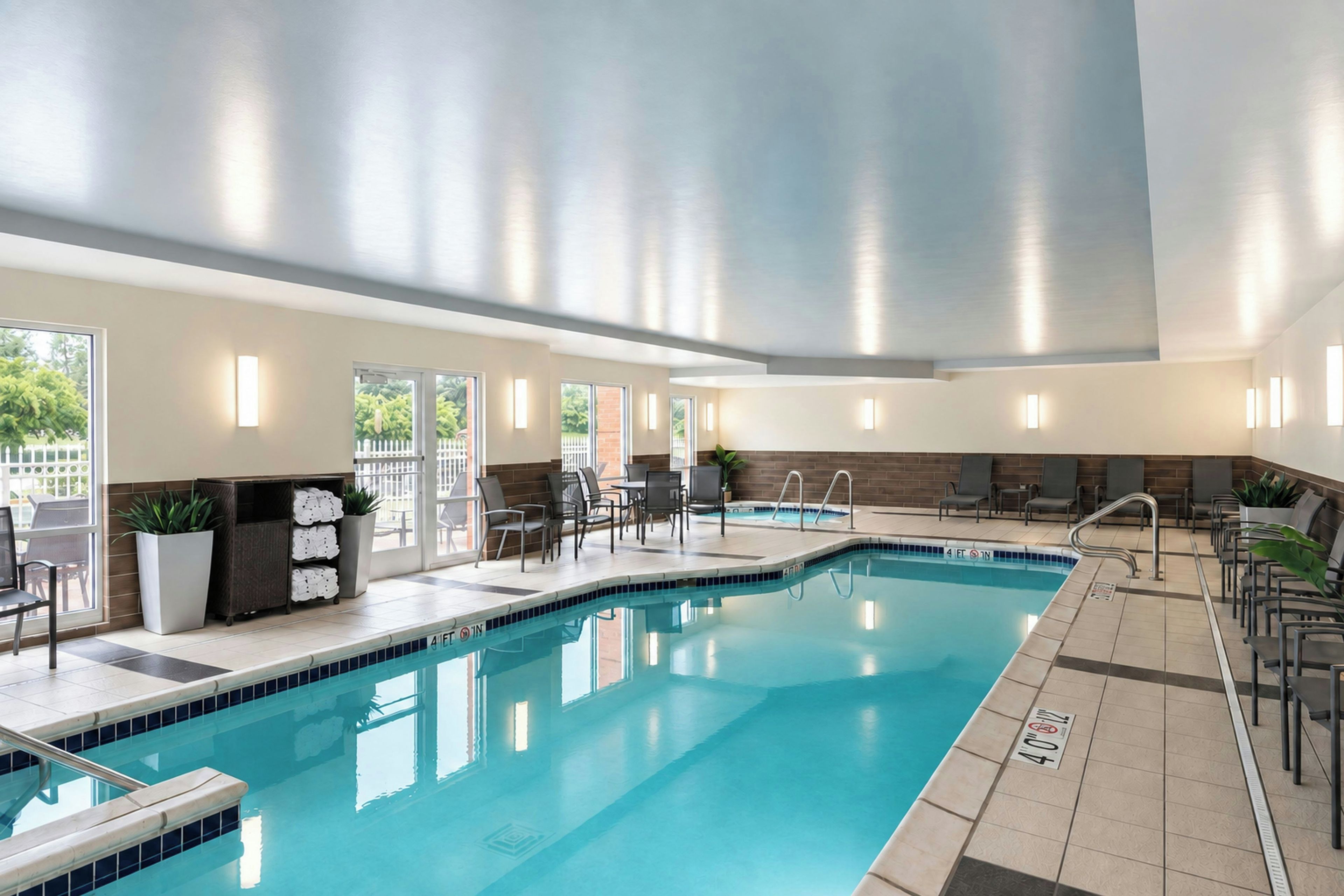 A swimming pool with a tiled edge, a lounge area with chairs and tables, and a view of the outdoors through large windows.