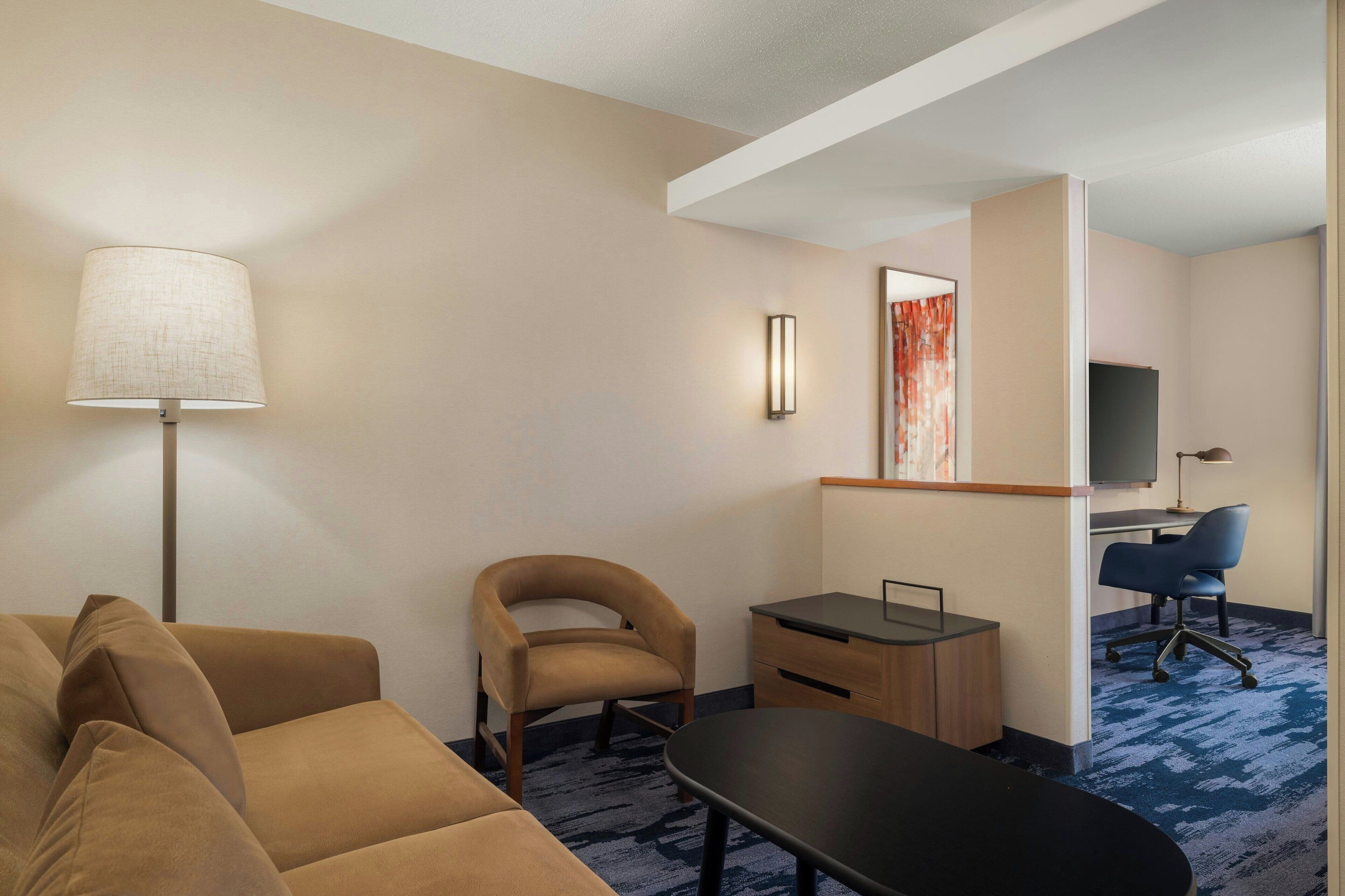 A hotel room with a sofa, a desk, a chair, a lamp, a TV, and a carpeted floor.