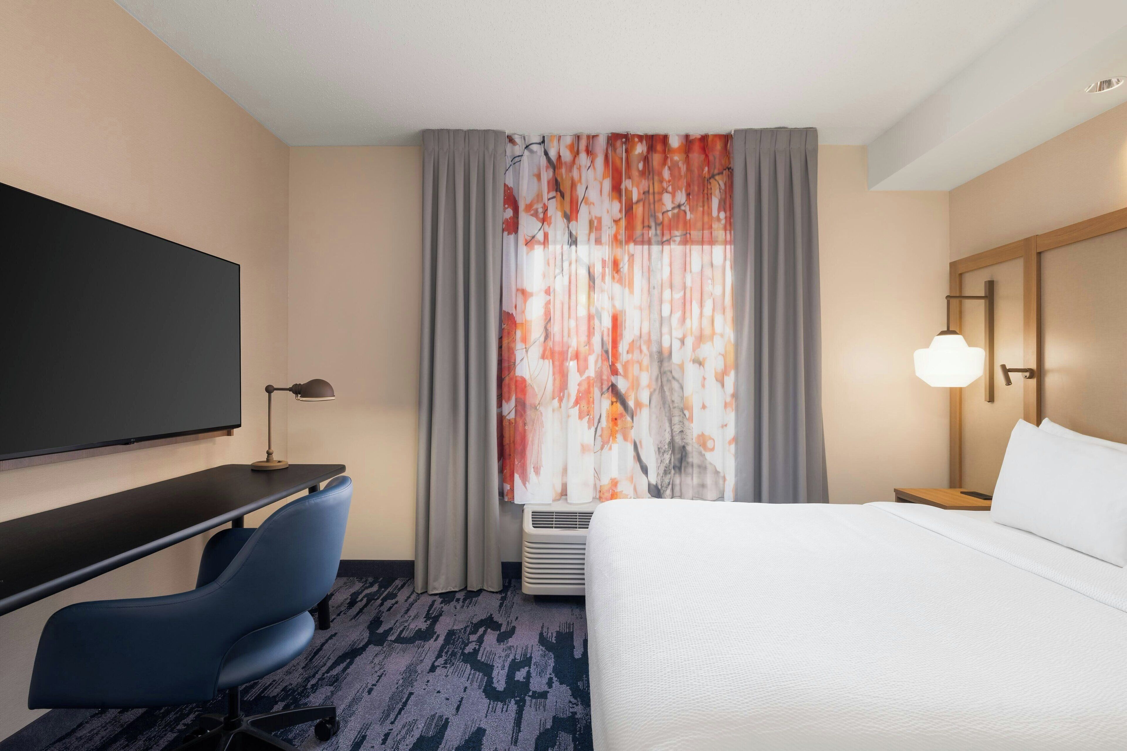 A hotel room with a bed, a desk with a chair, a flat-screen TV, a lamp, and a window with curtains.