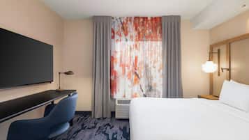 A hotel room with a bed, a desk with a chair, a flat-screen TV, a lamp, and a window with curtains.