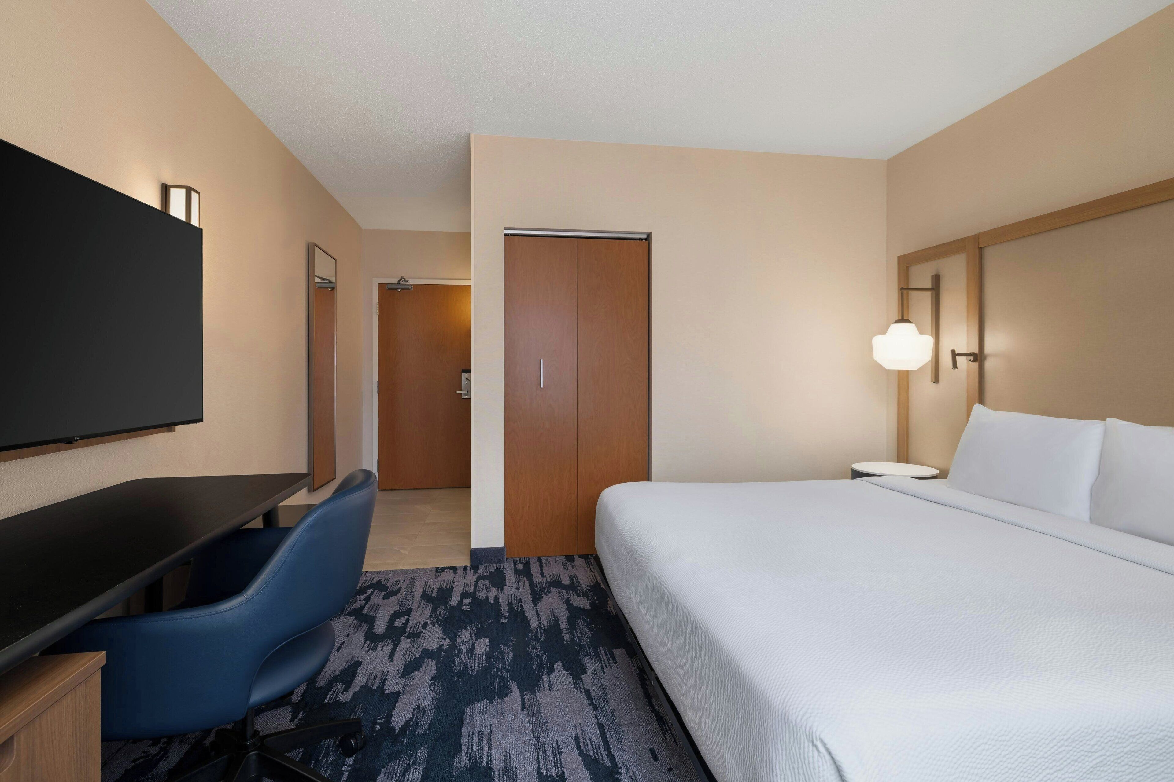 A hotel room with a bed, a desk with a chair, a television, and a door.