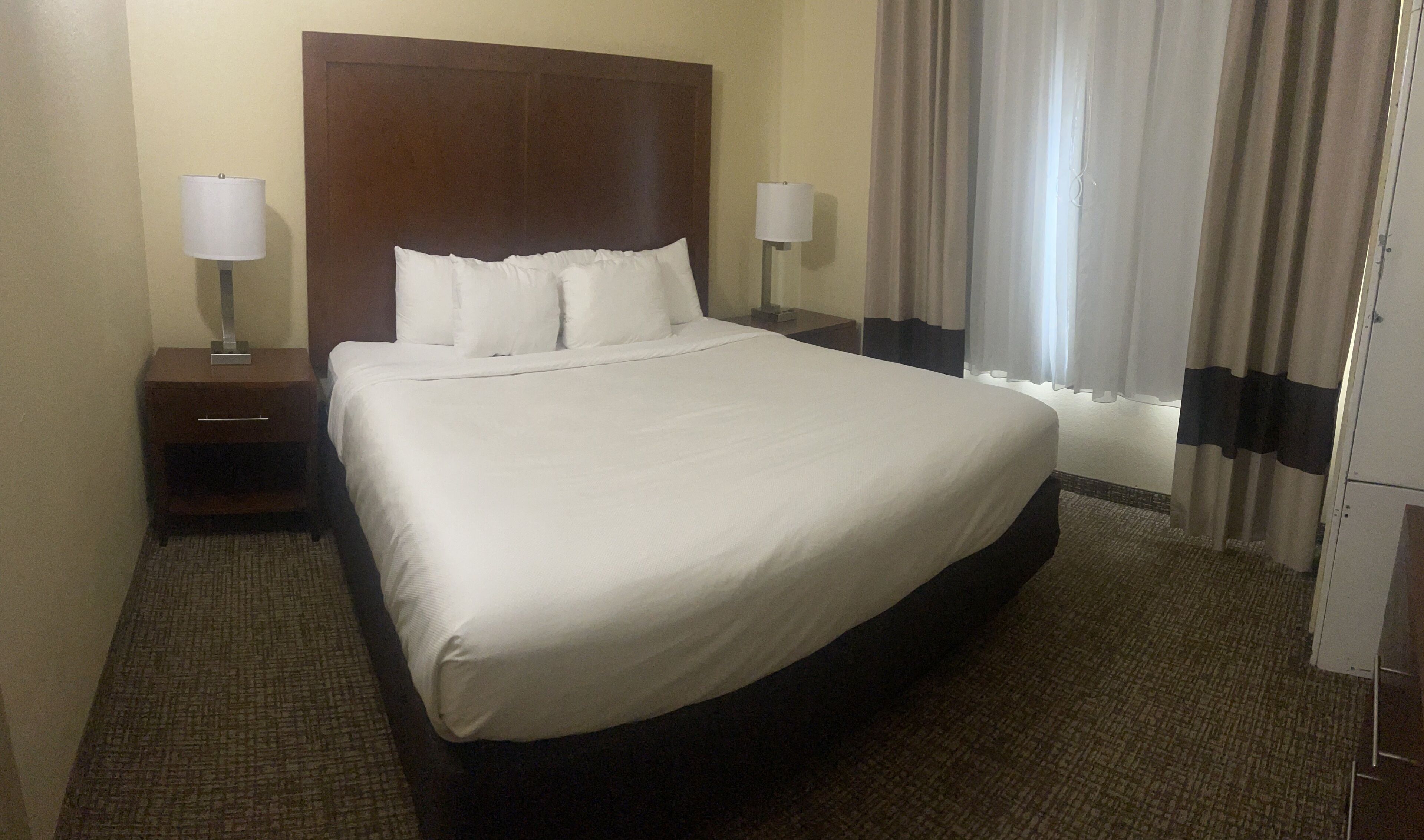 Habitación de hotel con una cama grande, dos mesitas de noche con lámparas, un cabecero y un ventanal con cortinas.