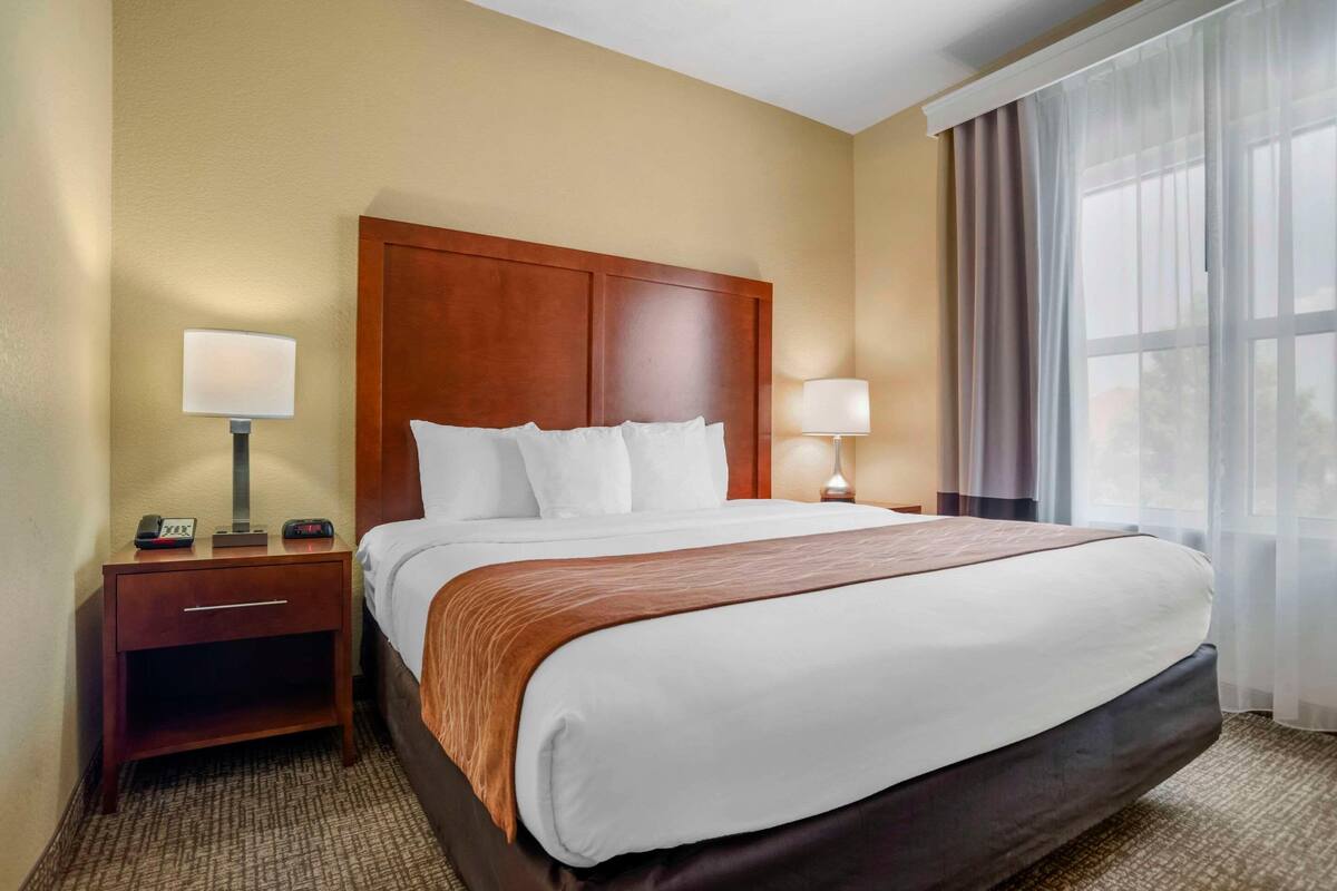 A hotel room with a large bed, two bedside tables with lamps, a wooden headboard, and a window with curtains.