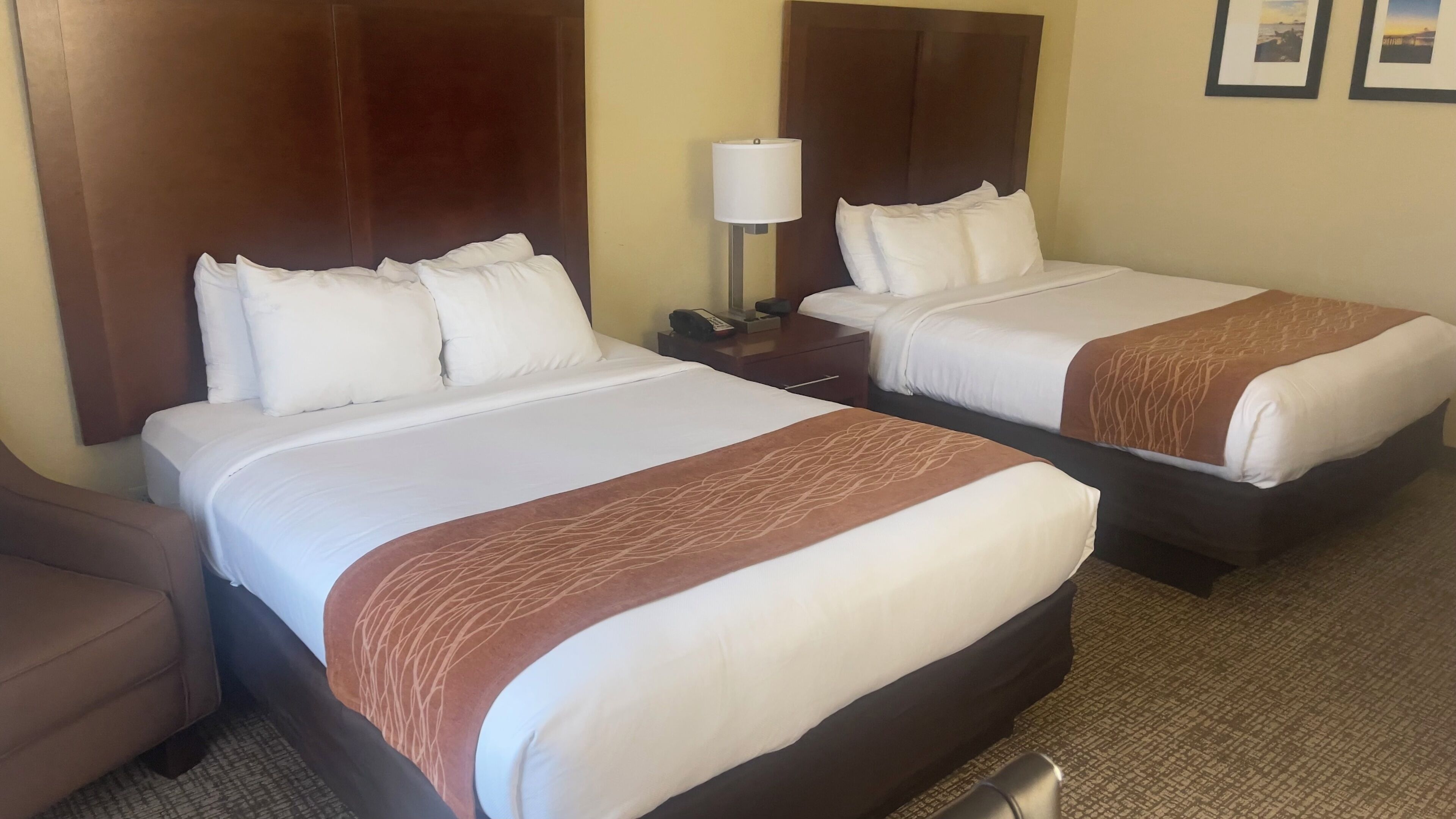 A hotel room with two beds, a nightstand with a lamp, and a chair.