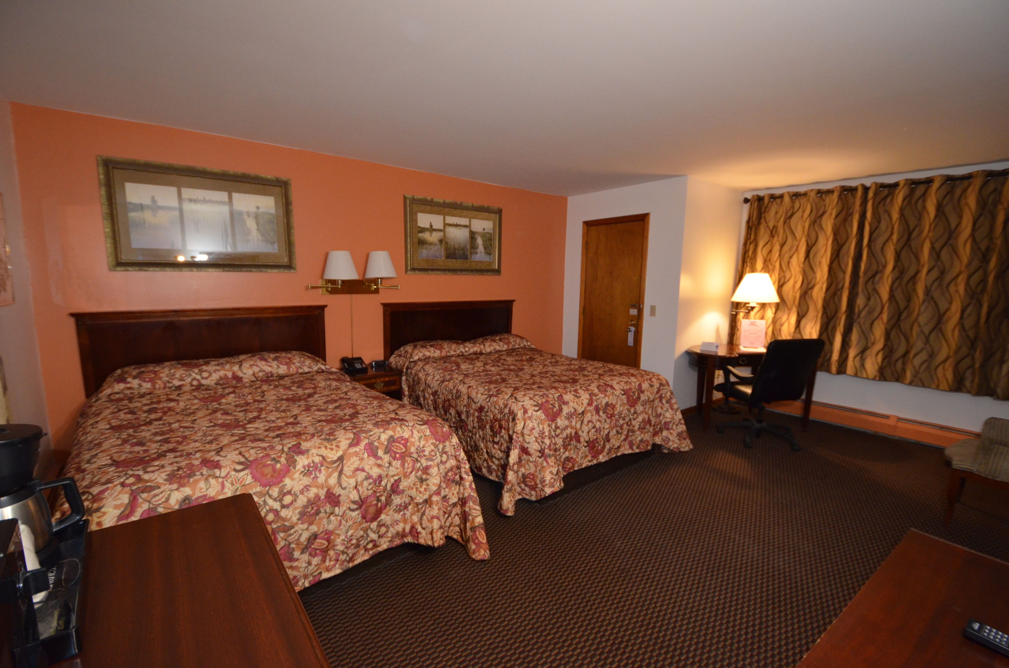 Quarto de hotel com duas camas, uma escrivaninha, uma cadeira, um abajur e uma porta.