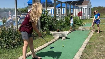 Un parcours de mini-golf avec une zone d’entraînement au putting verdoyante, une petite maison surmontée d’un drapeau rouge et un ciel d’un bleu limpide.
