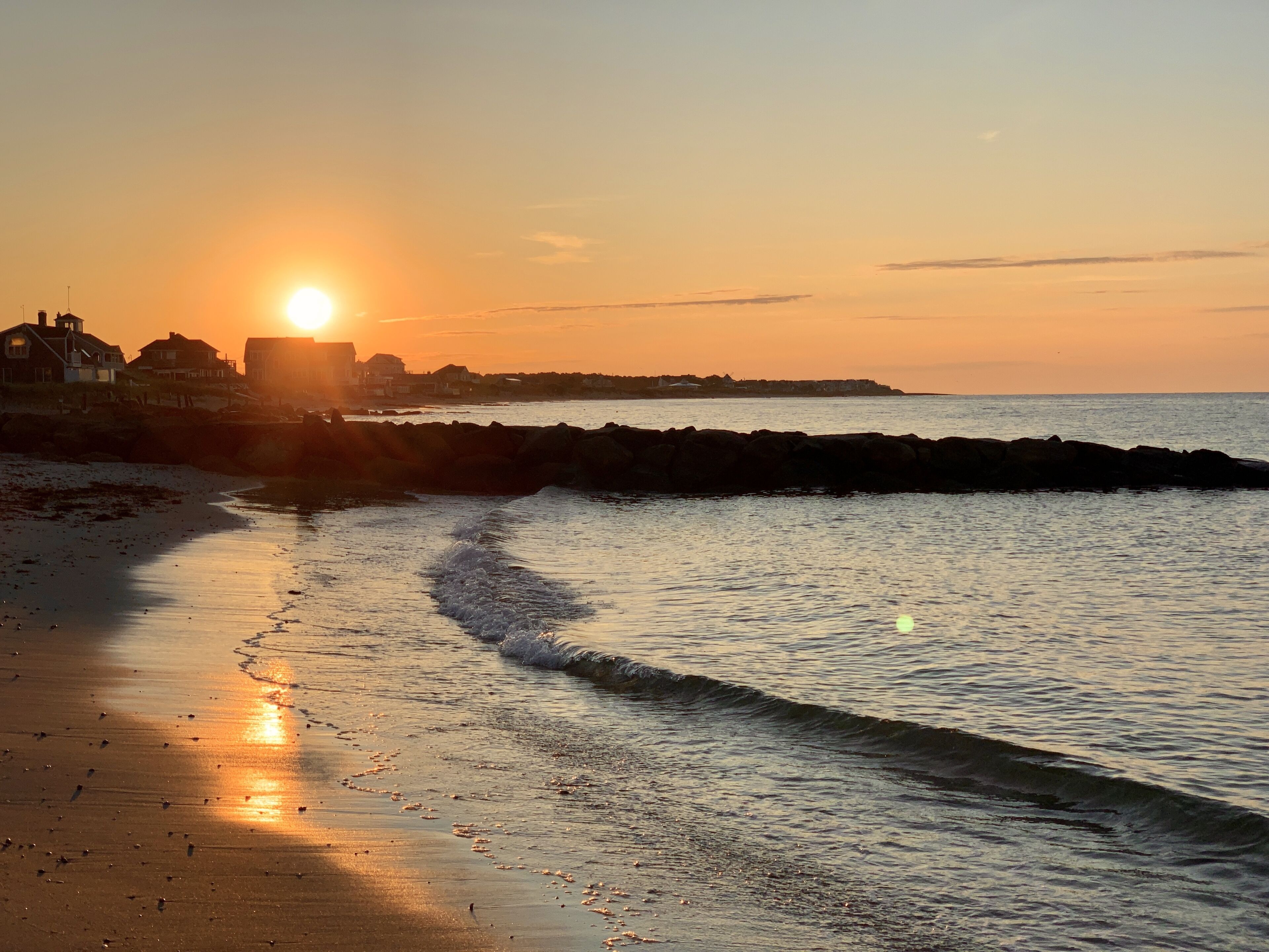 Escena de playa con el sol poniéndose en el horizonte, olas suaves lamiendo la orilla y edificios al fondo.