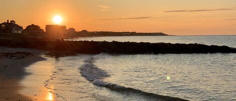 Escena de playa con el sol poniéndose en el horizonte, olas suaves lamiendo la orilla y edificios al fondo.