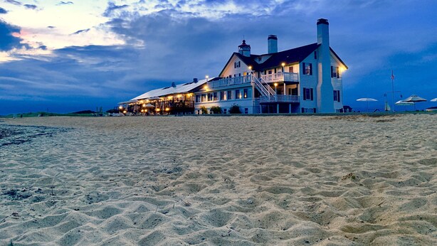 Un hotel grande frente a la playa, con varios pisos, un balcón destacado y múltiples ventanas.