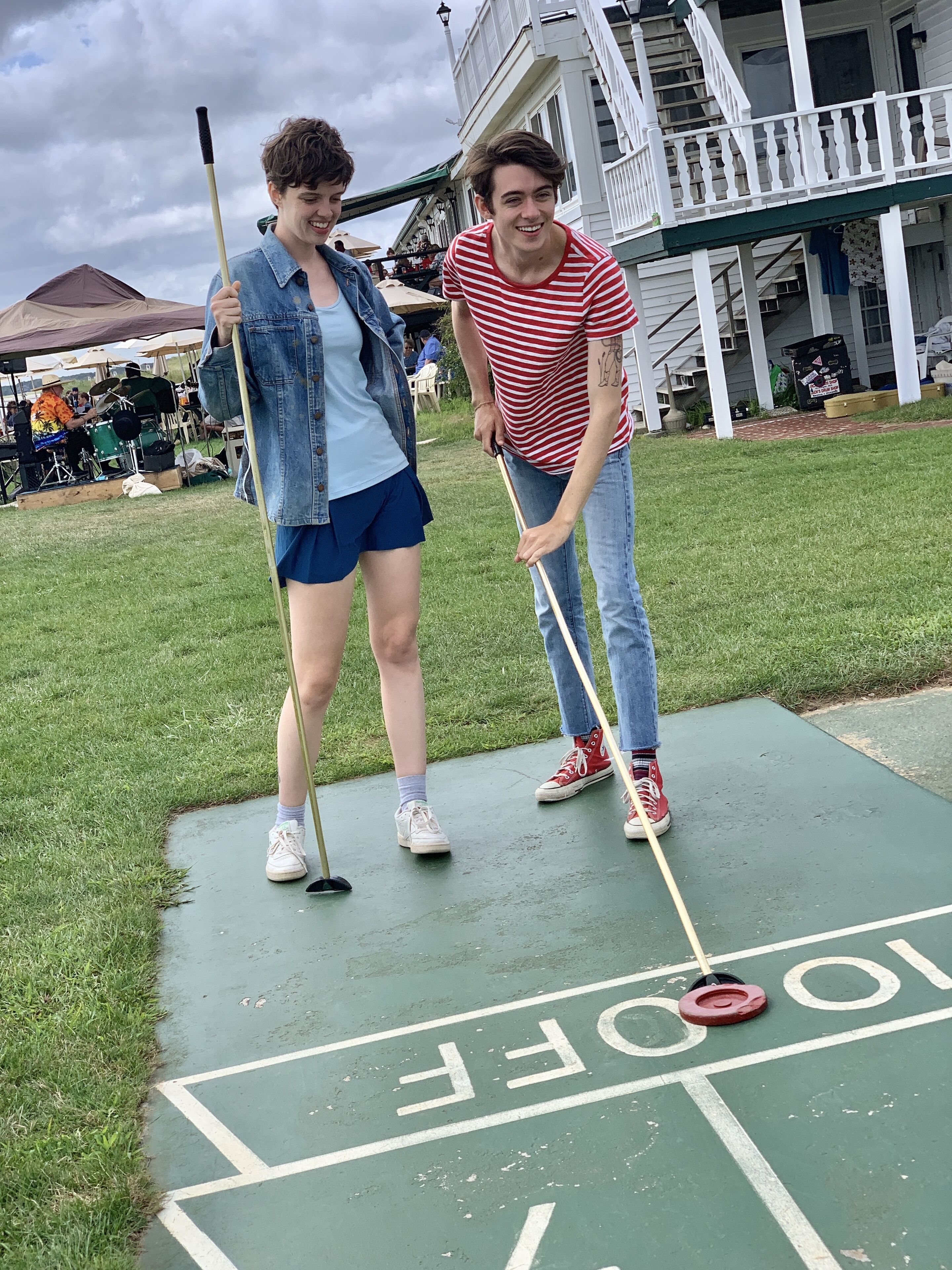Two people playing shuffleboard on a green court with a house in the background.