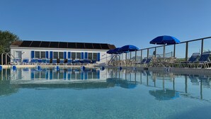 Una piscina cristalina con sillas y sombrillas azules, un edificio con contraventanas azules y un cielo despejado.