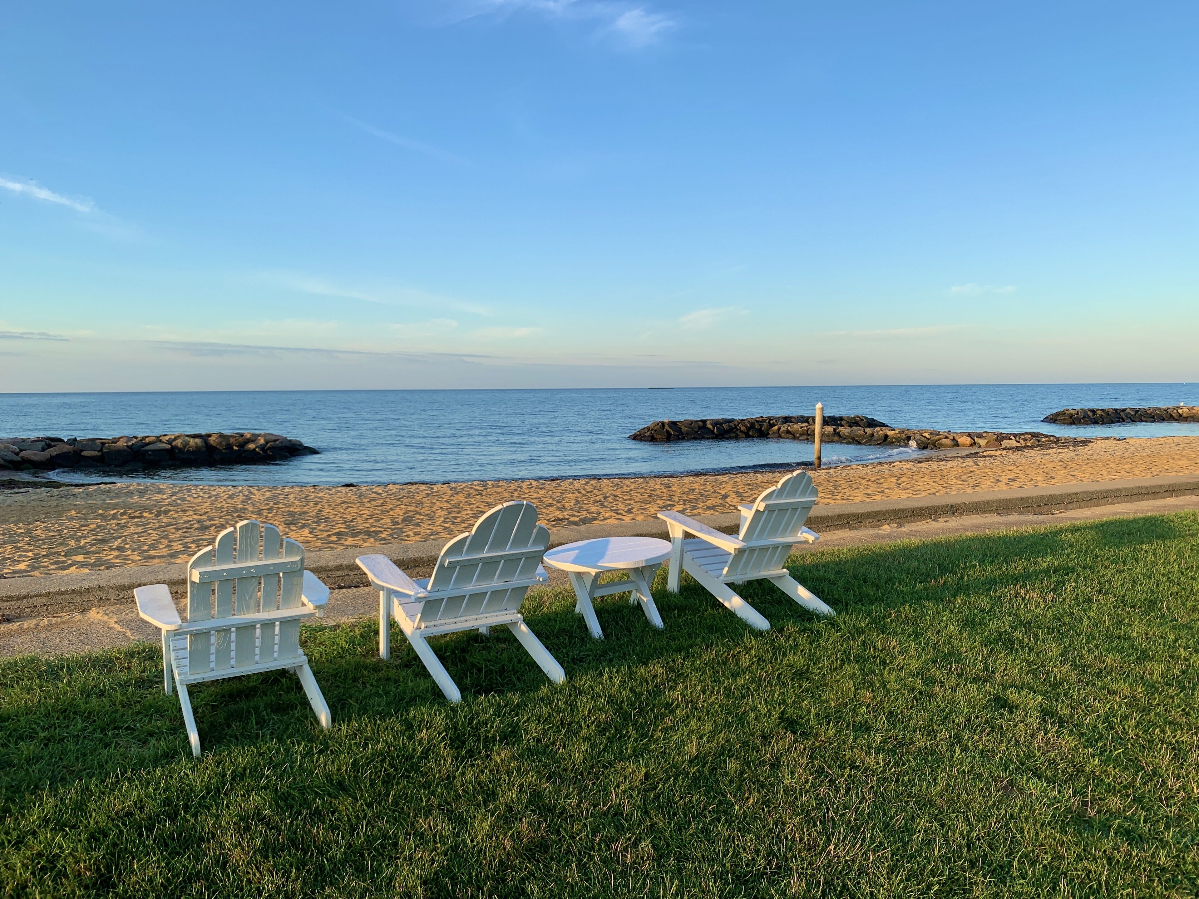 A beachside seating area with three Adirondack chairs and a small round table, overlooking a calm sea and rocky outcrops.
