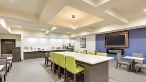 A modern hotel room with a large dining table, multiple chairs, a kitchen area, and a flat-screen TV mounted on the wall. - La Quinta Inn & Suites by Wyndham Tyler South (Tyler)