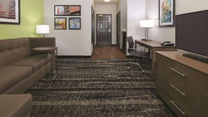 A hotel room with a brown sofa, a desk with a chair, a television, and a patterned carpet. - La Quinta Inn & Suites by Wyndham Tyler South (Tyler)