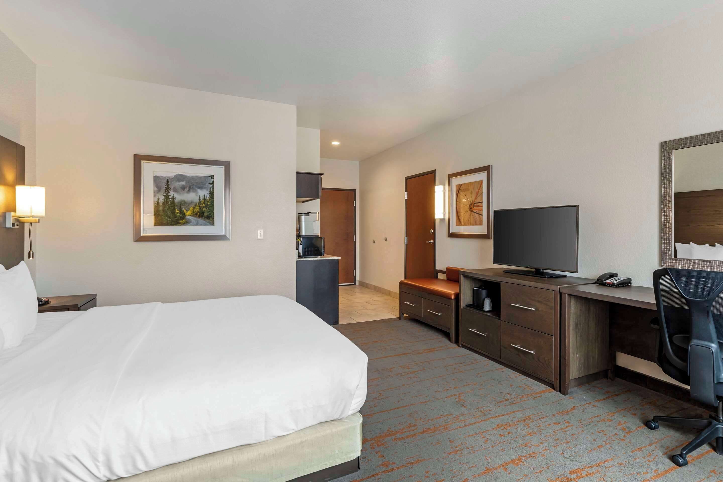 Habitación de hotel con una cama grande, un escritorio con una computadora, un televisor y un espejo. Hay cuadros enmarcados en las paredes y una lámpara en la mesita de noche.