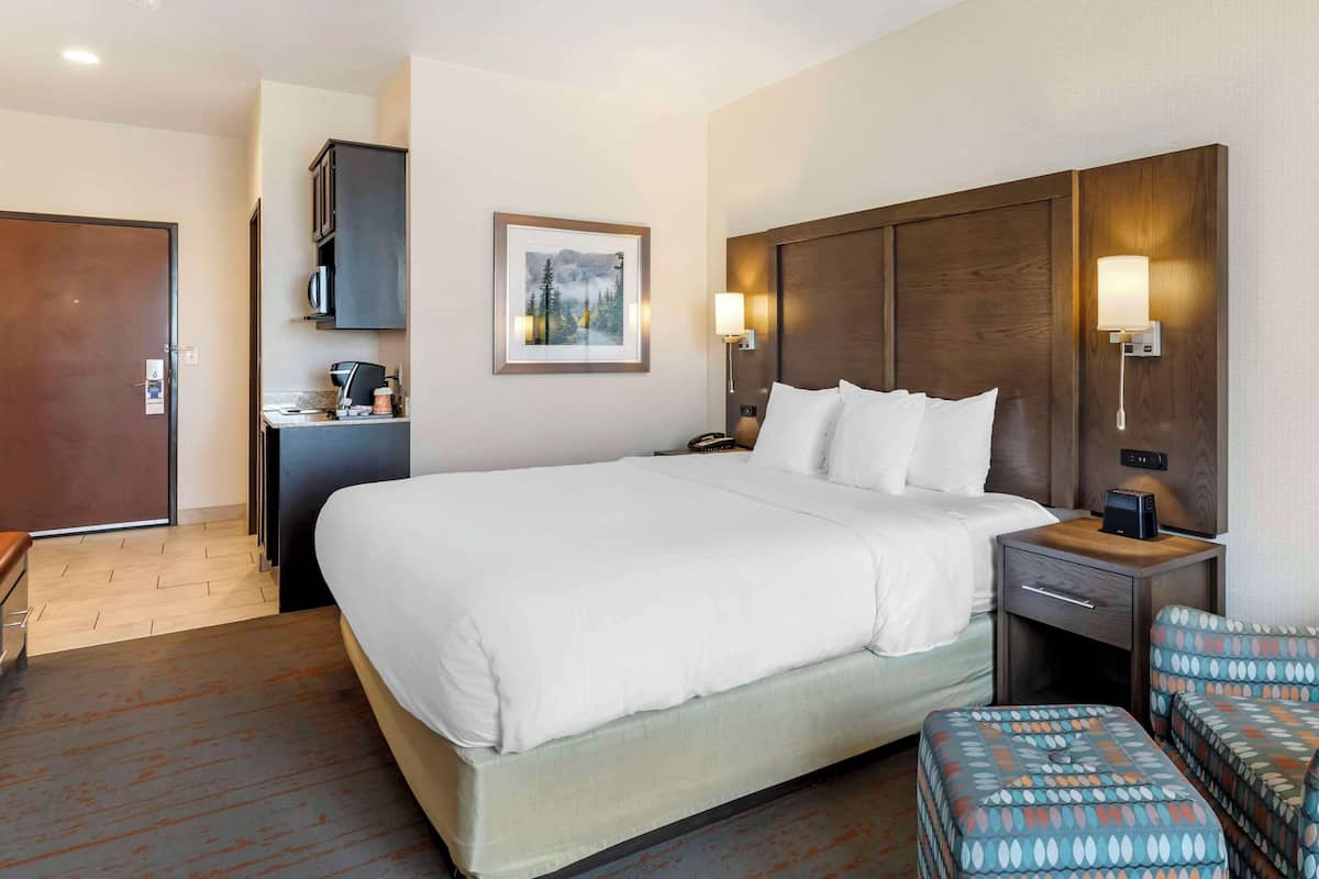 A hotel room with a large bed, a desk with a computer, a chair, and a small table with a lamp.